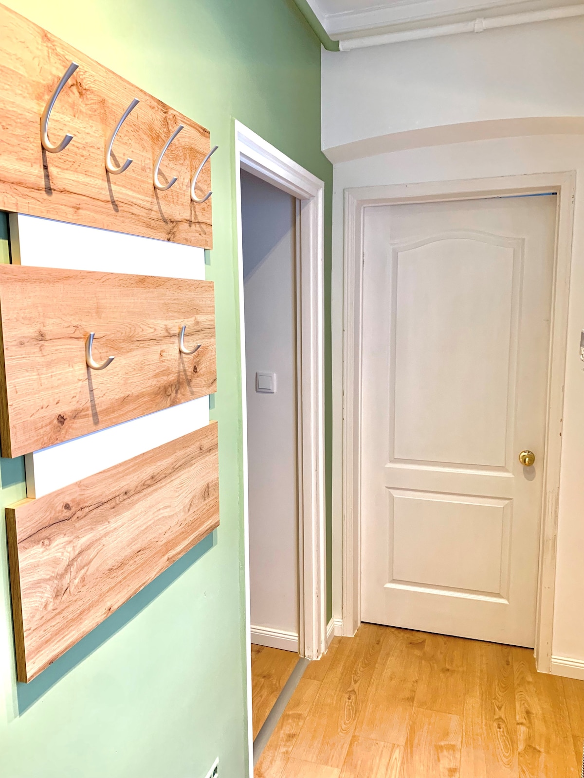 A hallway is shown with light green walls and wooden wall panels featuring several hooks for hanging items. A white door is visible, leading to another room, and warm wood flooring enhances the welcoming ambiance.