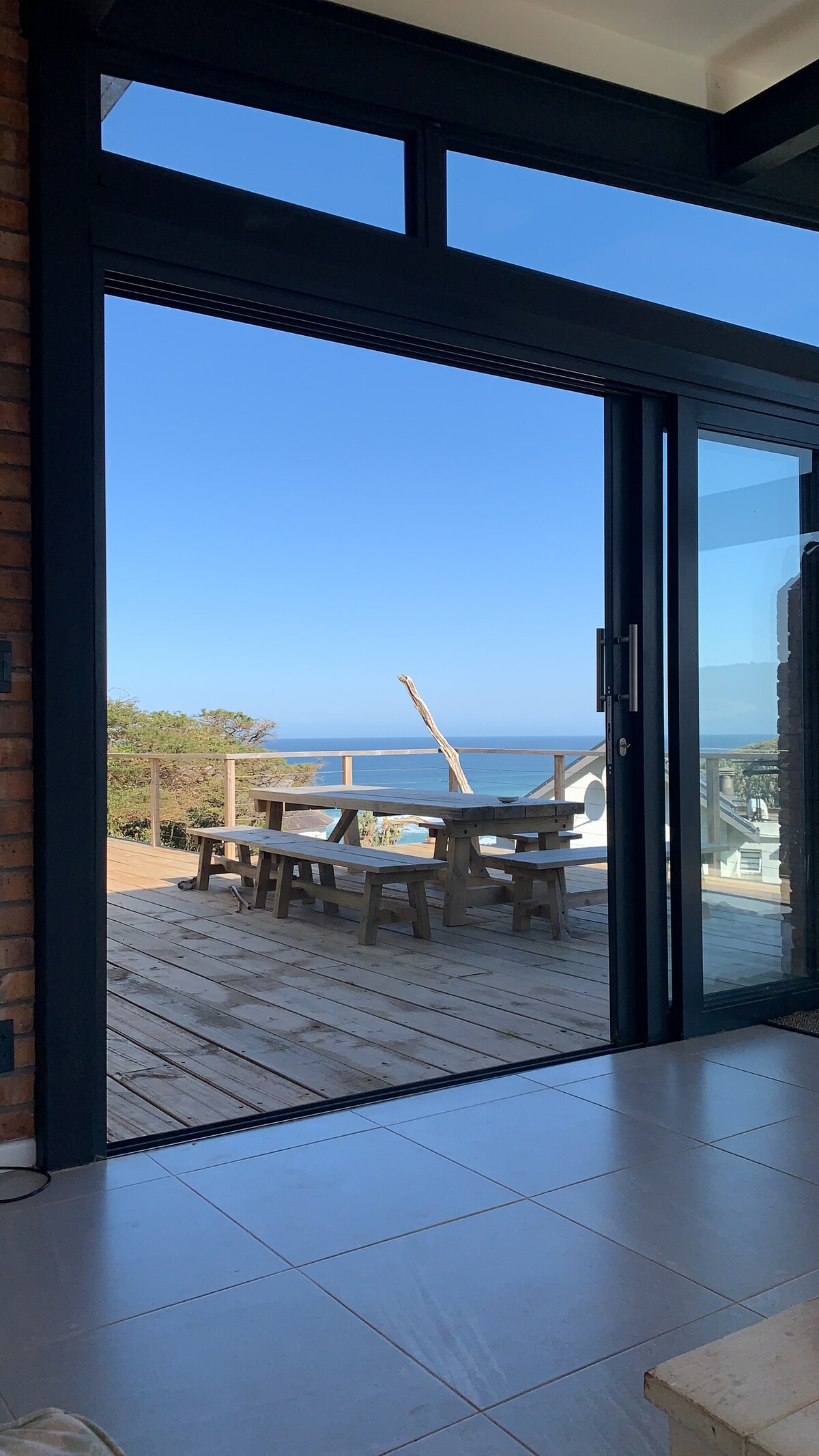 An expansive glass door opens to a scenic outdoor deck featuring a wooden table and benches, positioned against a backdrop of the ocean and clear blue sky. The inviting wooden deck is surrounded by natural landscaping.