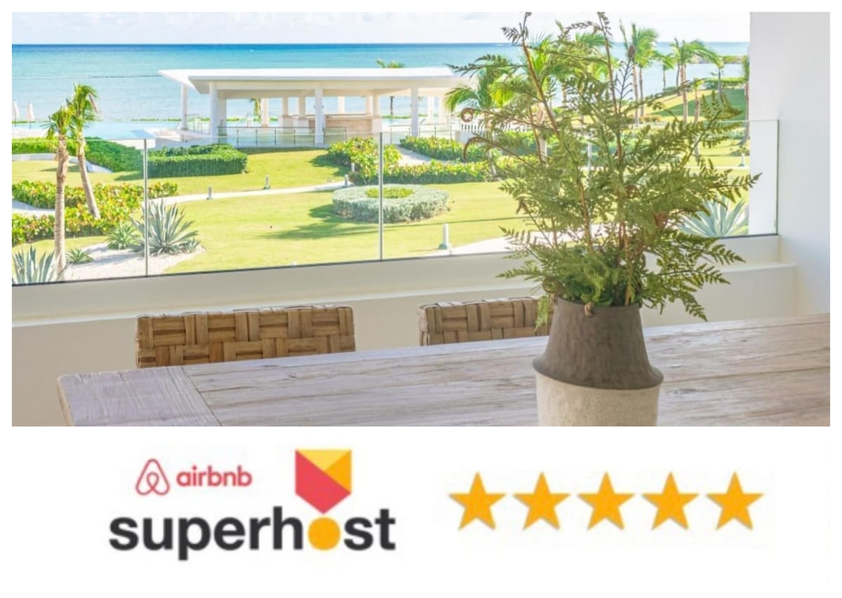 A serene view of the beach and ocean is framed by glass panels, revealing lush greenery and a well-kept lawn. A potted plant sits on a rustic wooden dining table, enhancing the tranquil atmosphere of the space.