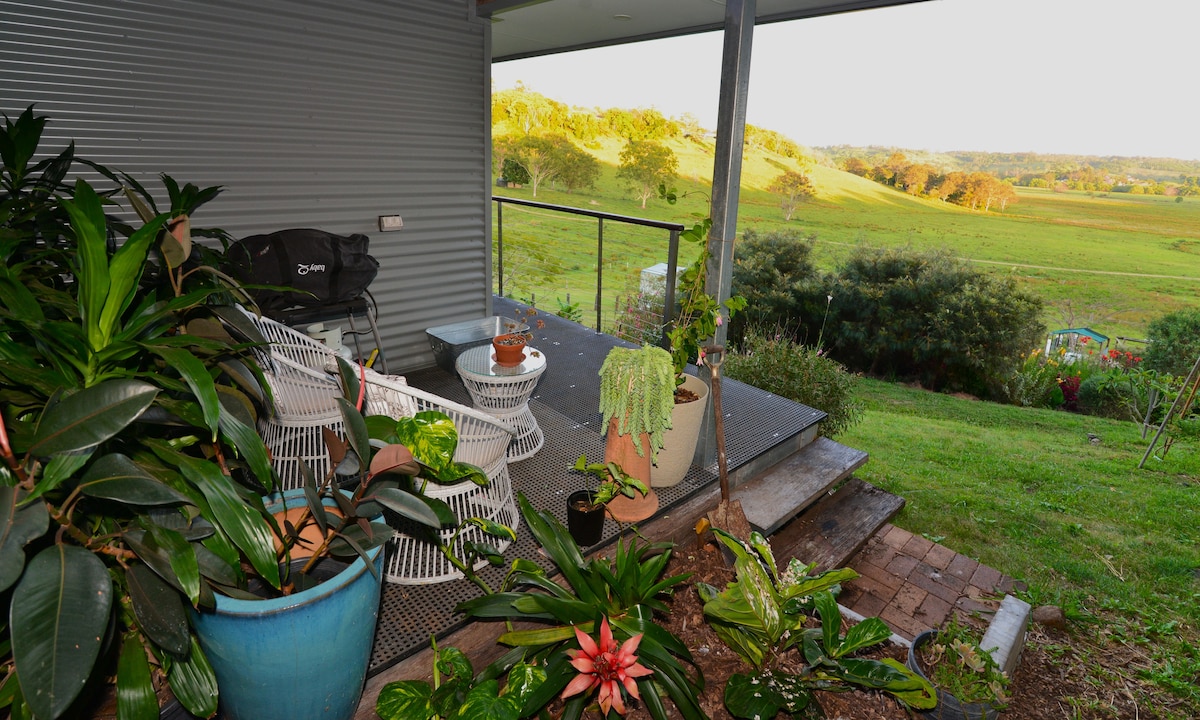 A wooden deck is observed with a few potted plants and comfortable seating arrangements. Lush greenery stretches out into the valley, creating a tranquil view. The area has a peaceful atmosphere, complemented by gentle natural surroundings.