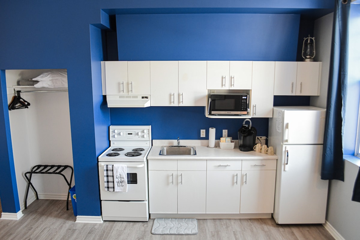 A compact kitchen area features modern white cabinetry against a vibrant blue wall. Essential appliances, including a stove, microwave, and refrigerator, are present. A sink is situated between the appliances, with kitchen utensils displayed neatly. A small closet is visible in the corner, offering additional storage.