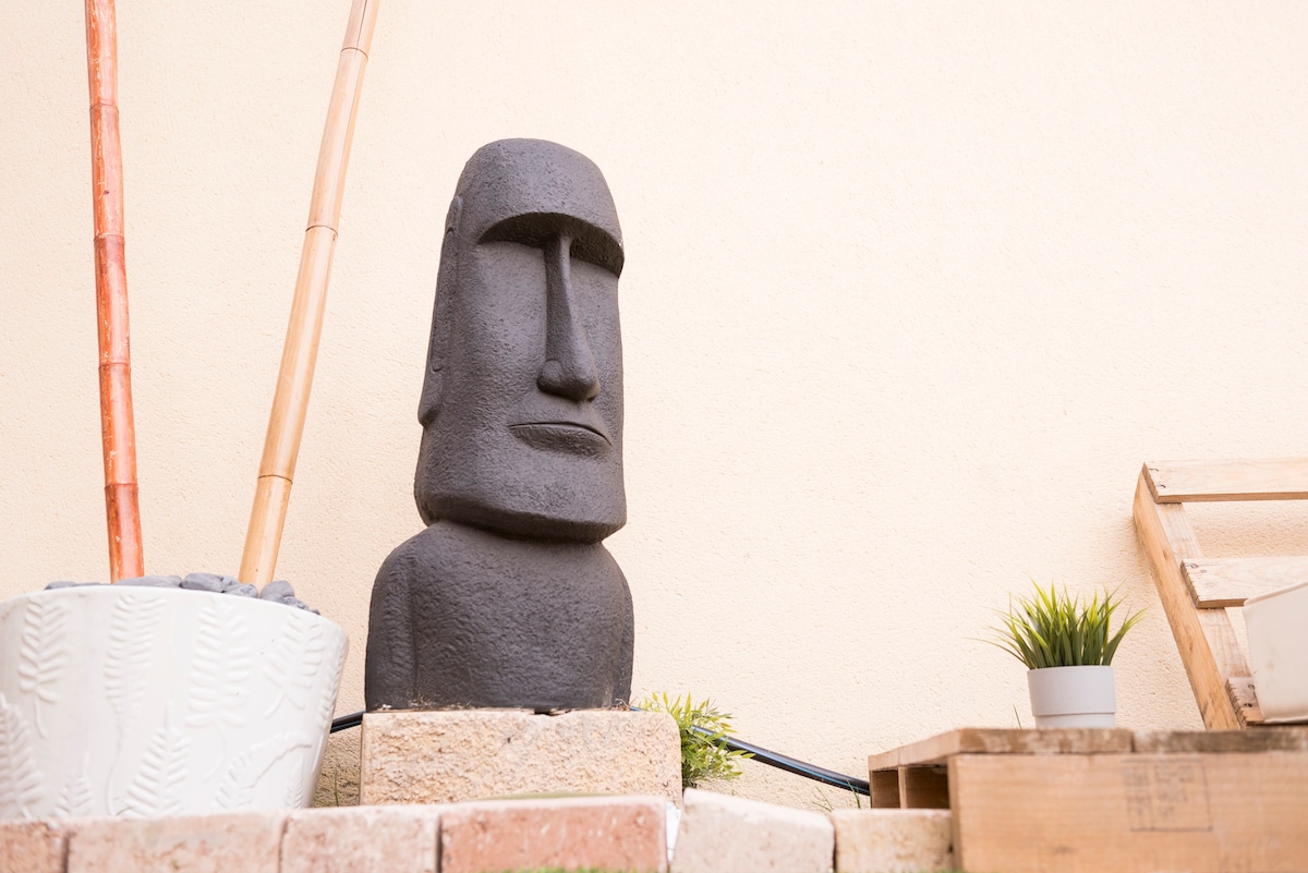 An intriguing Moai statue is prominently displayed, adding a touch of character to the outdoor area. Surrounding elements include a potted plant, bamboo sticks, and wooden crates, creating a relaxed and unique atmosphere.