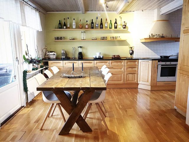 A kitchen space features wooden cabinetry and a long dining table with white chairs. Shelves above display various bottles and kitchen items. Natural light filters in through large windows, highlighting the wooden flooring and creating an open atmosphere.