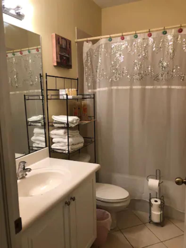 A shared bathroom features a white vanity with a sink and a large mirror above. A shower curtain with decorative elements is present, along with neatly stacked towels on a black metal rack. Essential toiletries and rolls of toilet paper are visible, alongside a small waste bin on the floor.