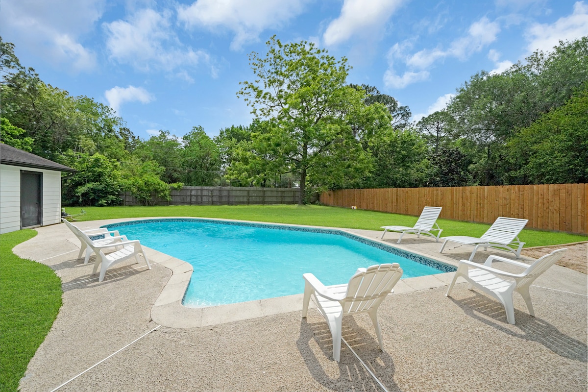 An outdoor pool area is showcased, featuring clear blue water surrounded by a textured concrete deck. Several white lounge chairs are positioned around the pool, and lush greenery forms a natural backdrop, adding to the serene environment.
