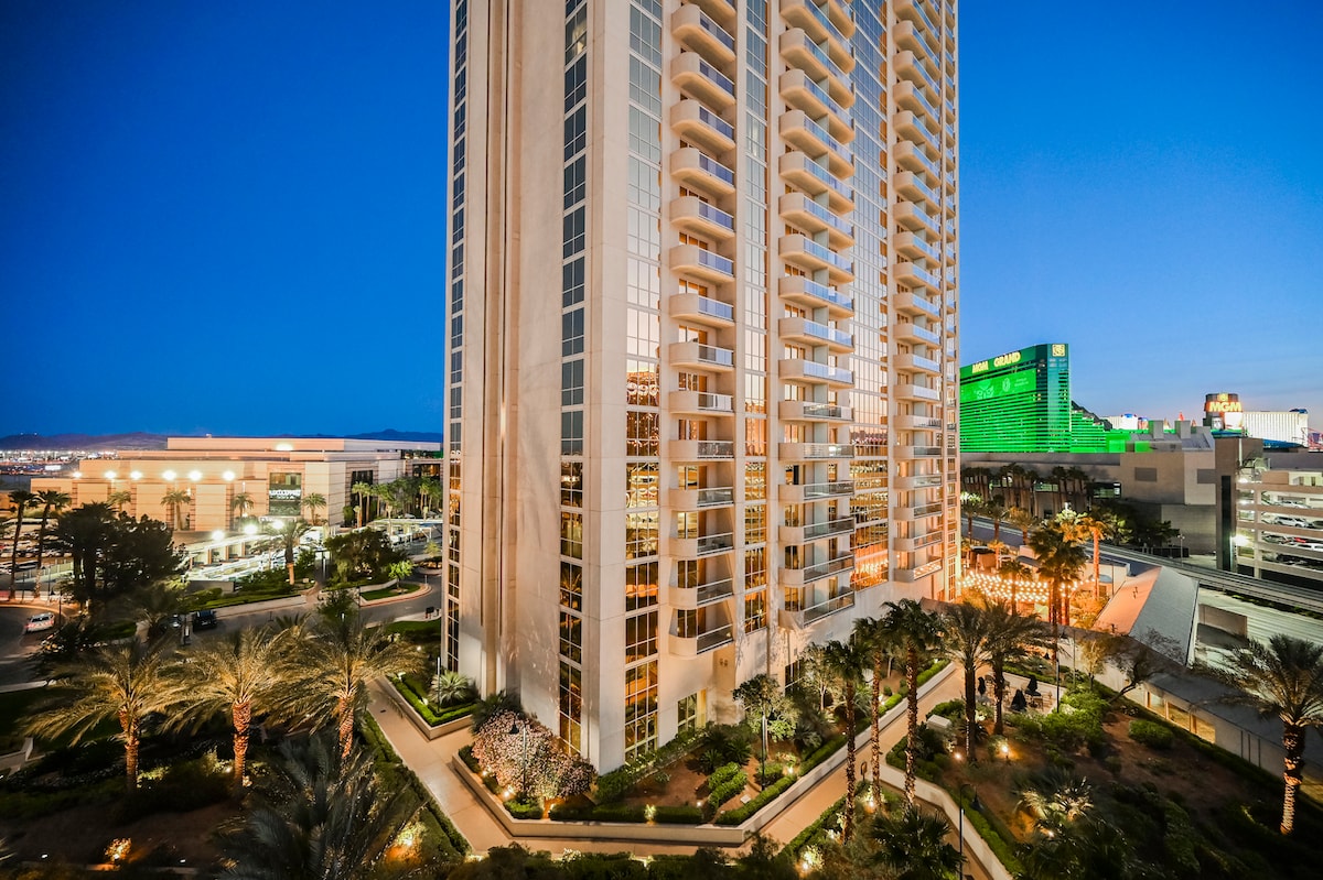 A tall, modern building with extensive glass exterior reflects the evening sky. Palm trees and illuminated landscaping surround the base, providing a welcoming entrance. The Las Vegas Strip is visible in the background, showcasing vibrant neon lights and nearby attractions.