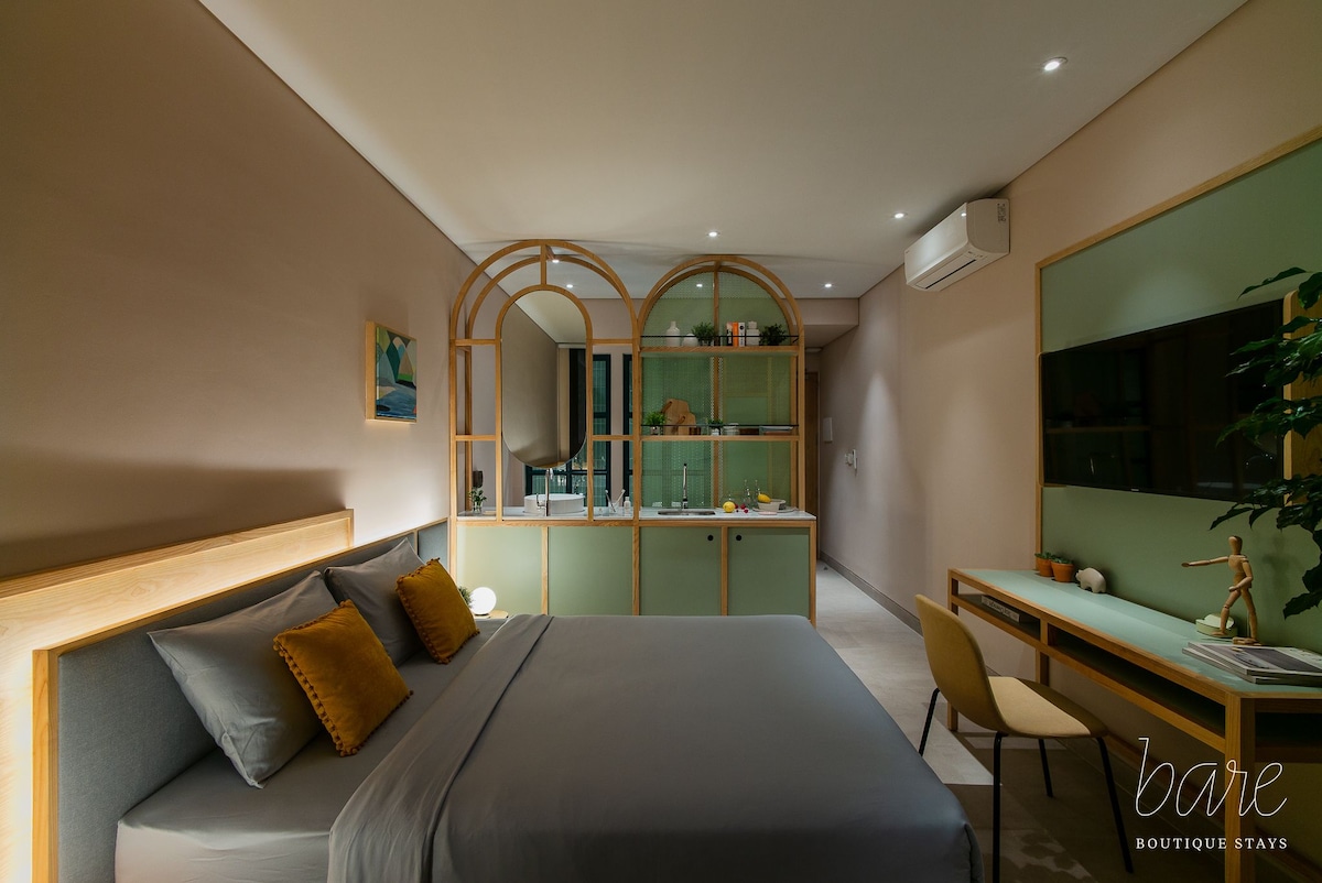 A modern bedroom is presented with a spacious bed dressed in soft gray linens. A wooden accent wall complements the light green shelving unit, which displays decorative items. A small desk and chair are positioned next to a wall-mounted television, enhancing the functional space.