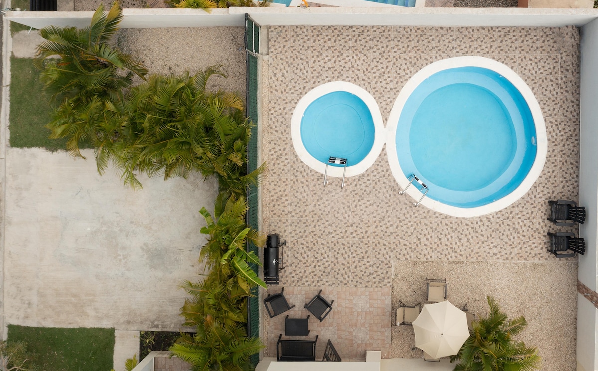 An overhead view showcases a private pool area designed with two interconnected pools, surrounded by a patterned stone patio. Several lounge chairs are arranged nearby, with a shaded table and umbrella positioned for comfort. Lush greenery adds a natural touch to the outdoor space.