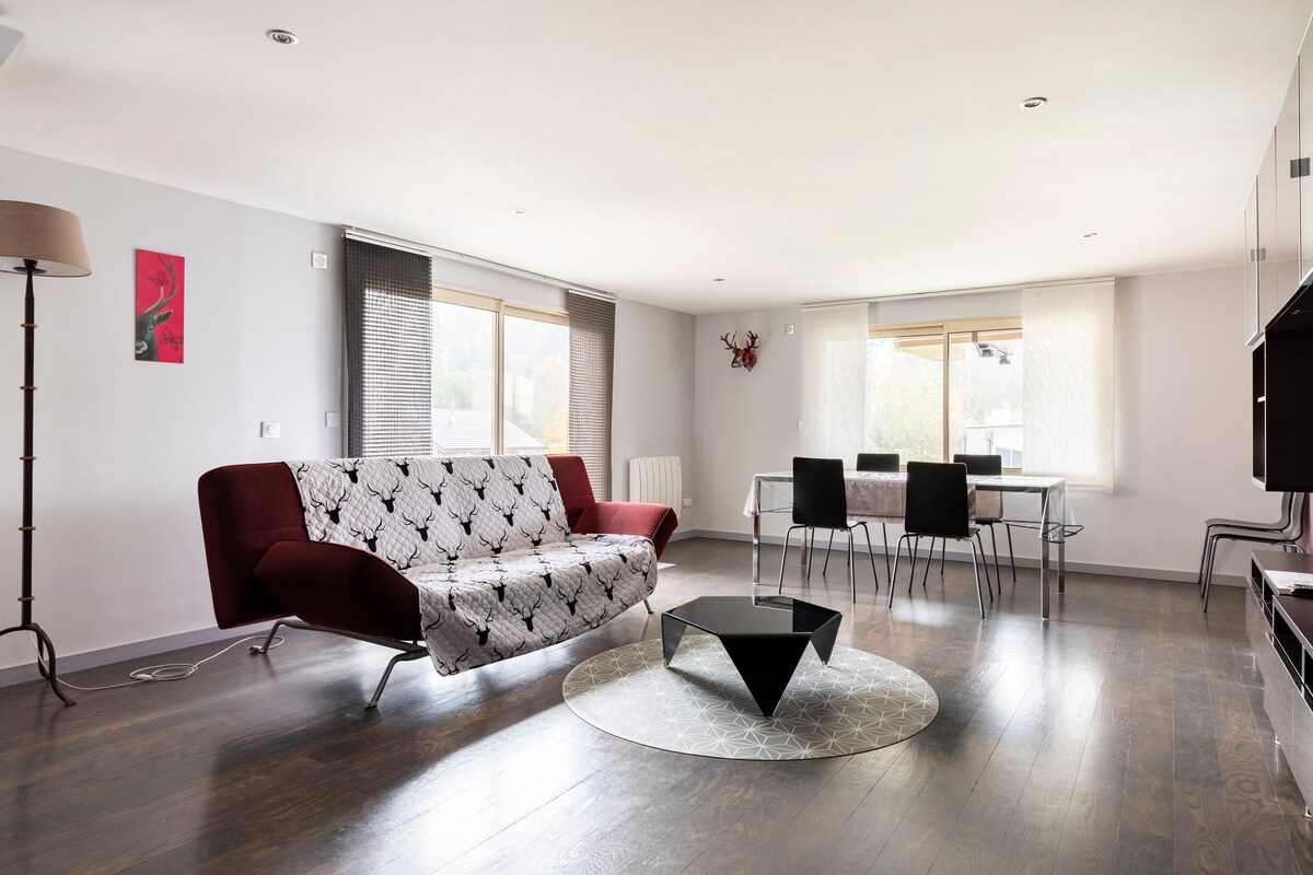A spacious living area is furnished with a comfortable red sofa and a modern glass dining table surrounded by sleek metal chairs. Natural light filters in through large windows, illuminating the room's wooden floor and minimalist design. A decorative rug anchors the seating area.
