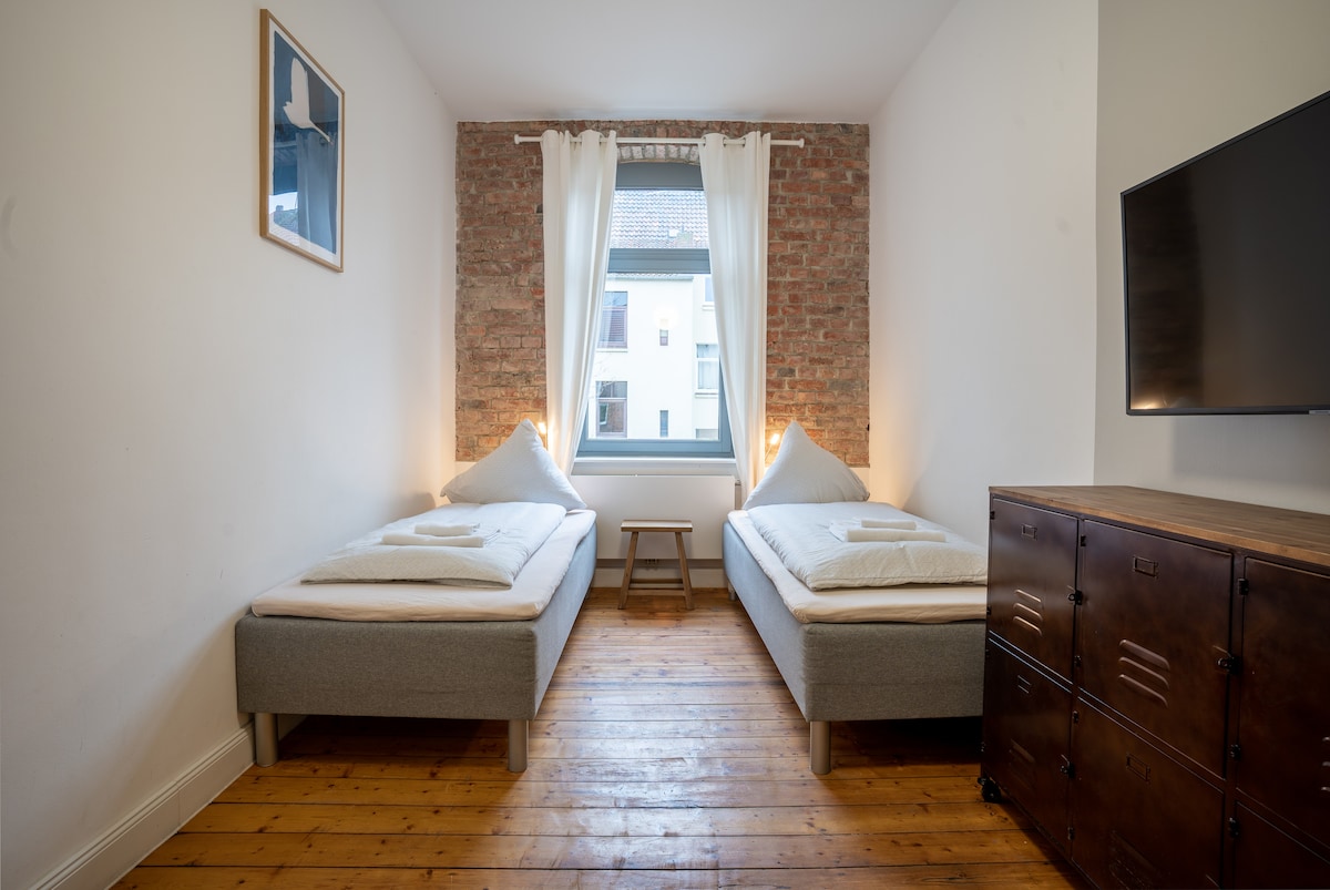 The room features two individual beds with soft linens arranged neatly. A window allows natural light to fill the space, framed by light curtains. A small wooden table sits between the beds, and a dark wooden dresser is positioned against the wall.