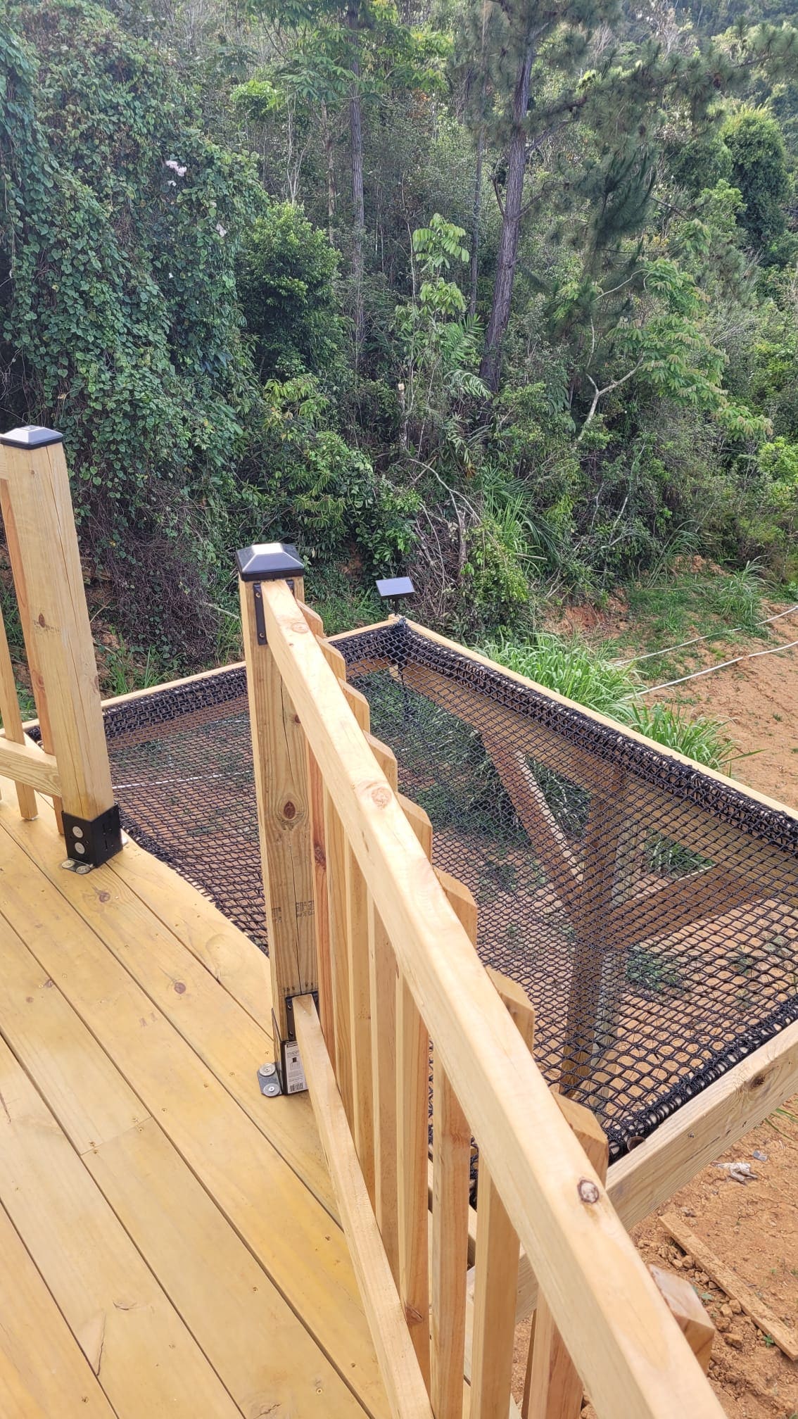 A wooden deck railing leads to a netted area that extends over a natural landscape filled with greenery. The surrounding trees provide a sense of seclusion, while the sturdy construction invites relaxation and an appreciation of the outdoor environment.