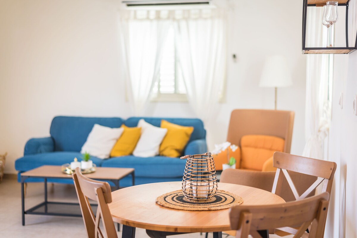 A cozy living space features a blue sofa with colorful throw pillows and an orange armchair. A wooden dining table is set in the center, adorned with a woven placemat and a decorative lantern. Natural light filters through sheer curtains, brightening the room.
