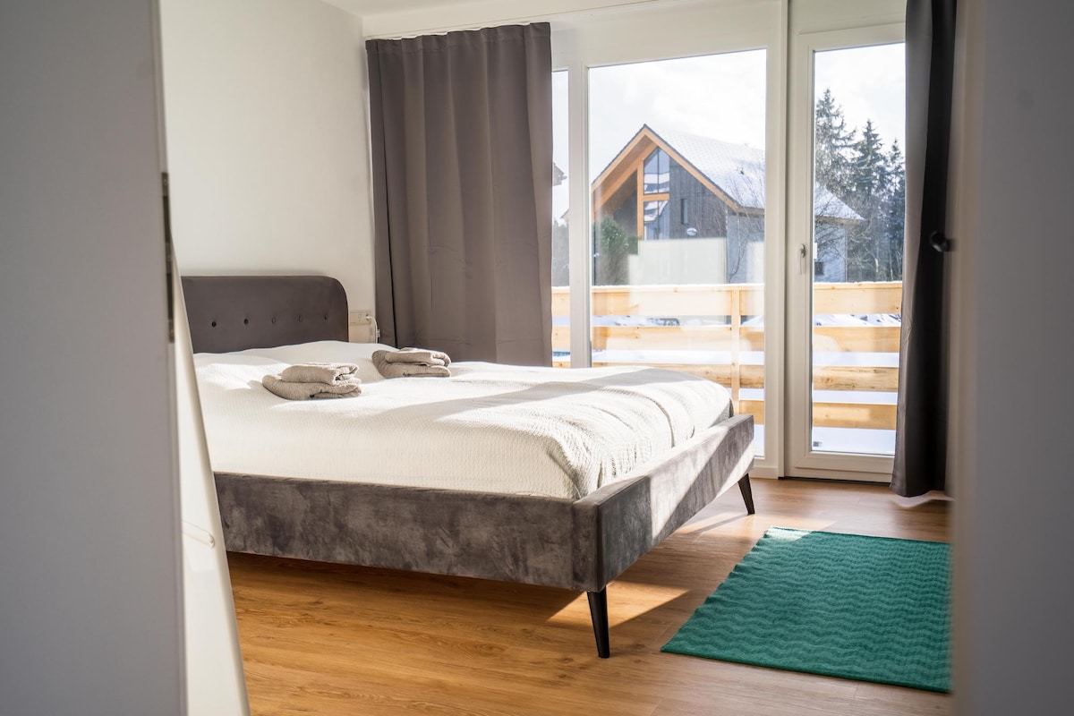 A spacious bedroom is highlighted by large glass doors that open to a balcony, allowing natural light to fill the room. A comfortable bed is dressed in light linens, with rolled towels placed neatly on top. The wooden floor is complemented by a green textured rug.