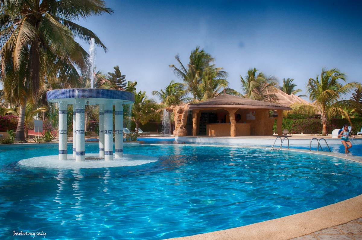 A large swimming pool is surrounded by palm trees, featuring an elegant fountain in the center. A covered seating area is visible to the right, providing a space for relaxation. Sun loungers are positioned nearby, emphasizing the inviting atmosphere of the outdoor area.
