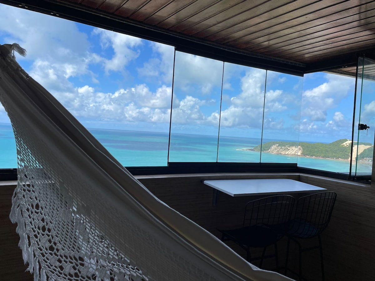 A hammock is suspended near large glass windows, providing an unobstructed view of the ocean and sky. A small table with two chairs is positioned nearby, allowing for enjoyment of the scenic surroundings.