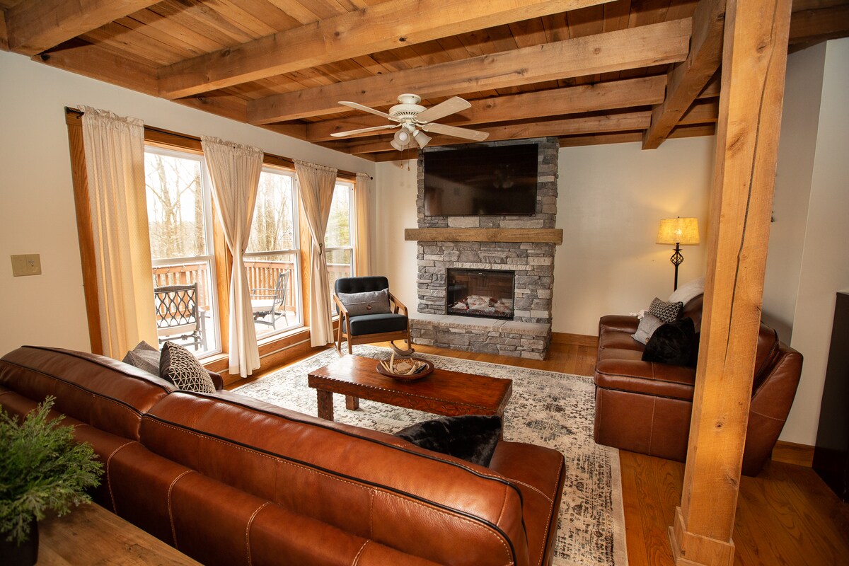 A welcoming living room features a stone fireplace and a large television mounted above. Natural light floods in through expansive windows framed with light curtains, revealing outdoor views. Comfortable leather sofas and a wooden coffee table create a cozy seating arrangement beneath exposed wooden beams.