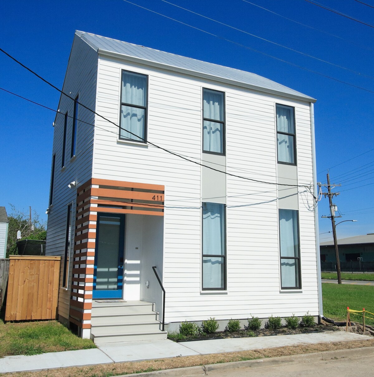 A modern cottage-style home is depicted, featuring a light-colored exterior with a slanted roof. Large windows allow ample natural light, and a contrasting entry door with a small porch adds a welcoming touch. Neatly arranged landscaping lines the walkway leading to the entrance.