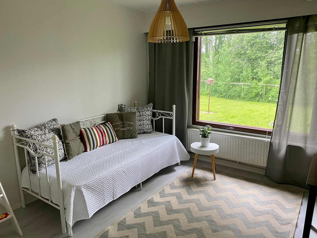 A bright living area features a white daybed adorned with patterned cushions, complemented by a small round table. A large window offers a view of the lush green outdoors, with sheer curtains allowing natural light to fill the space. A patterned rug adds warmth.
