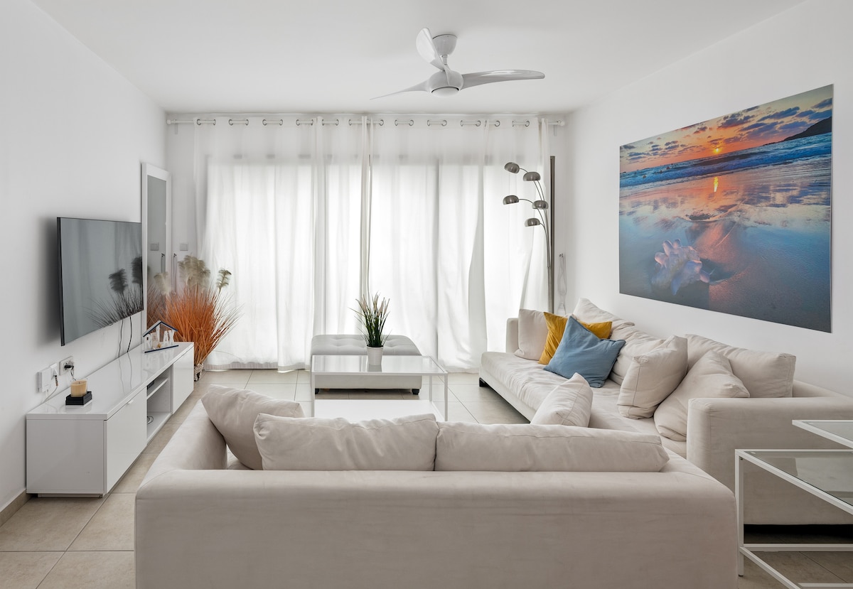 A spacious living room is filled with natural light, featuring large windows covered by sheer curtains. A cream-colored sectional sofa is accompanied by a white coffee table. A wall-mounted television and a piece of artwork depicting a beach sunset add to the room's comfort.