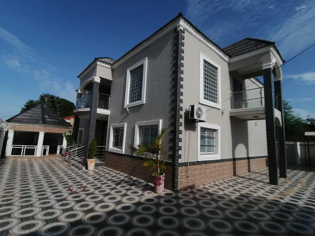 The exterior of the villa is showcased, featuring a two-story structure with a combination of light-colored walls and intricate decorative accents. A spacious tiled area surrounds the building, with potted plants adding a touch of greenery. Clear skies and ample sunlight illuminate the scene.