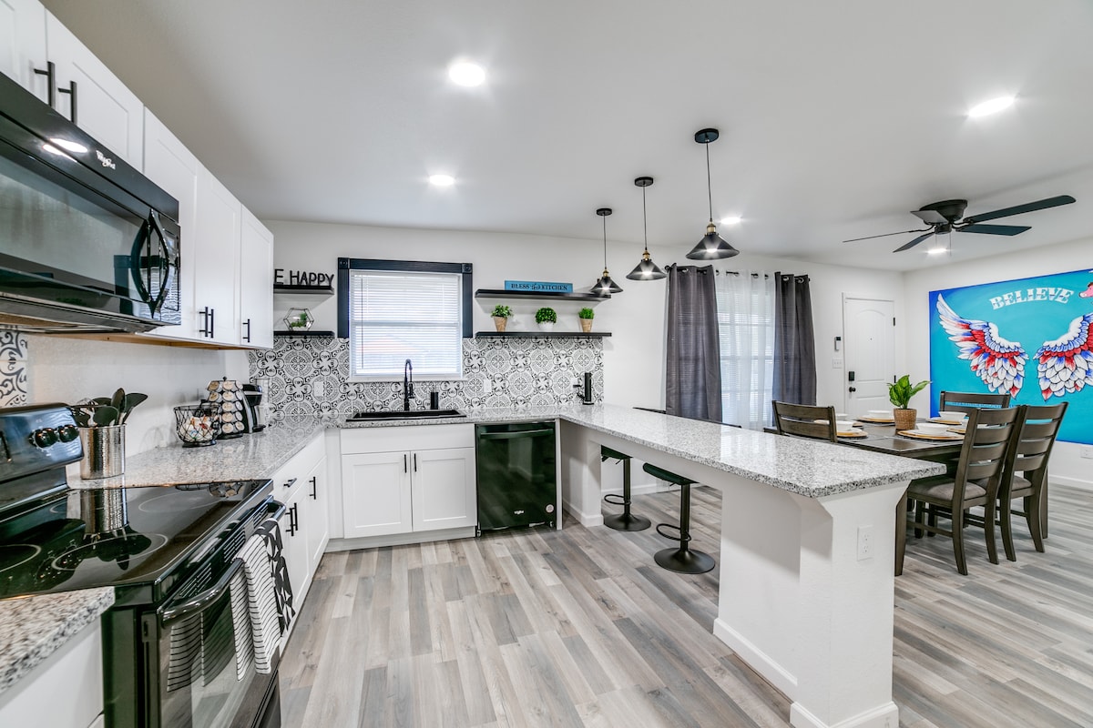 A modern kitchen and dining area are viewed in this image. The kitchen features stainless steel appliances and a spacious island with granite counters. Black pendant lights provide illumination over the dining table, while a bright mural decorates one wall.