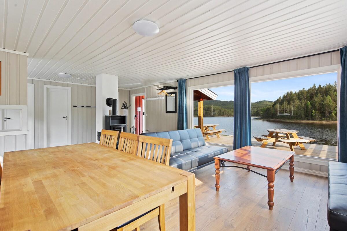 A spacious living area features a large wooden dining table, a comfortable seating arrangement, and large windows that offer views of the lake and surrounding forest. Natural light floods the room, creating a serene atmosphere.