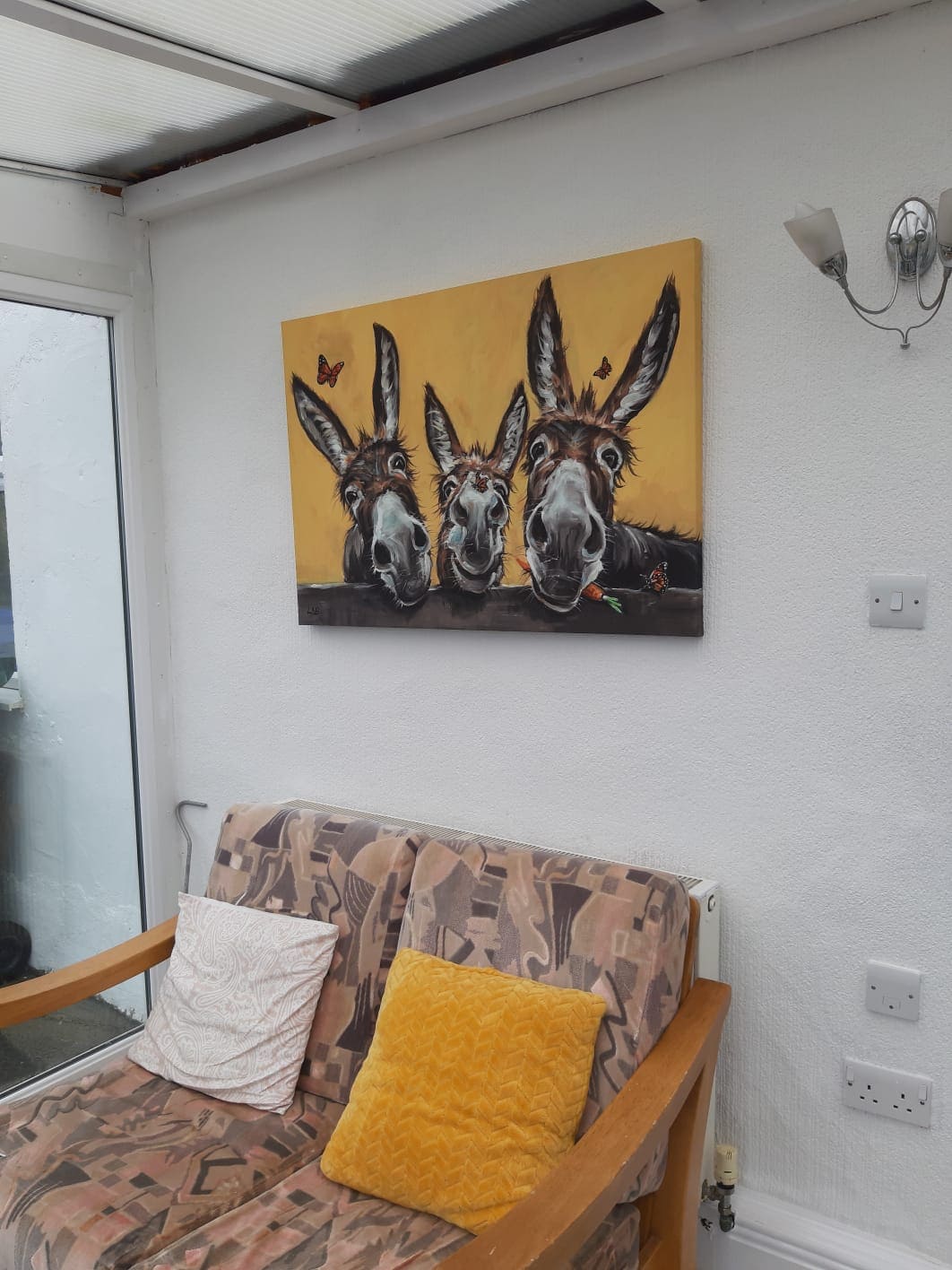 A seating area features a patterned couch accompanied by a yellow cushion. A whimsical painting of three donkeys adorns the wall, set against a bright yellow background. Natural light filters in through the nearby glass walls, creating a cheerful ambiance.