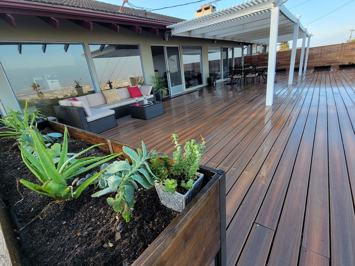 An expansive deck is showcased, featuring wooden flooring and comfortable outdoor seating arrangements. Succulent plants are neatly arranged in boxes along the edges, enhancing the natural atmosphere. Large glass doors allow access to the interior space, inviting light and open views of the surroundings.
