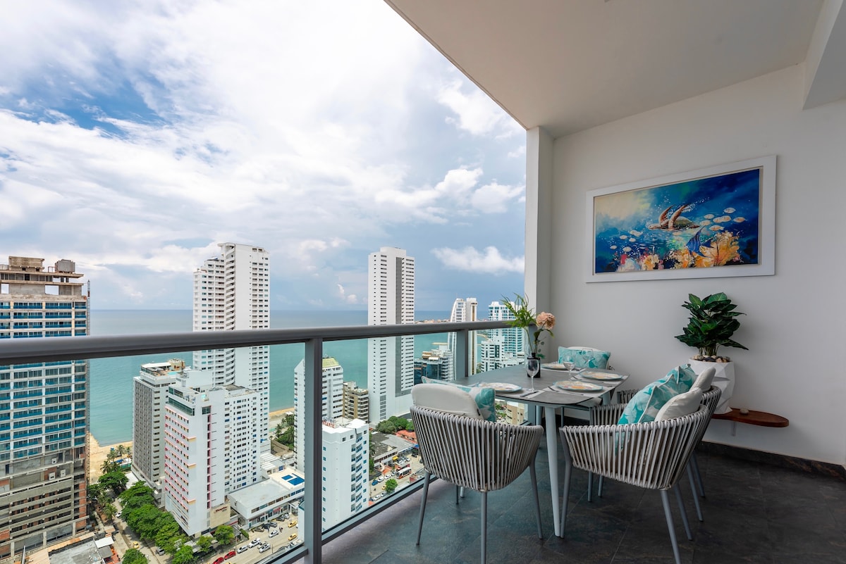 A balcony dining area offers scenic views of the Caribbean Sea and the skyline of the city. A round table is set with tableware, surrounded by four modern chairs. A vibrant piece of wall art and a potted plant add a touch of color to the space.