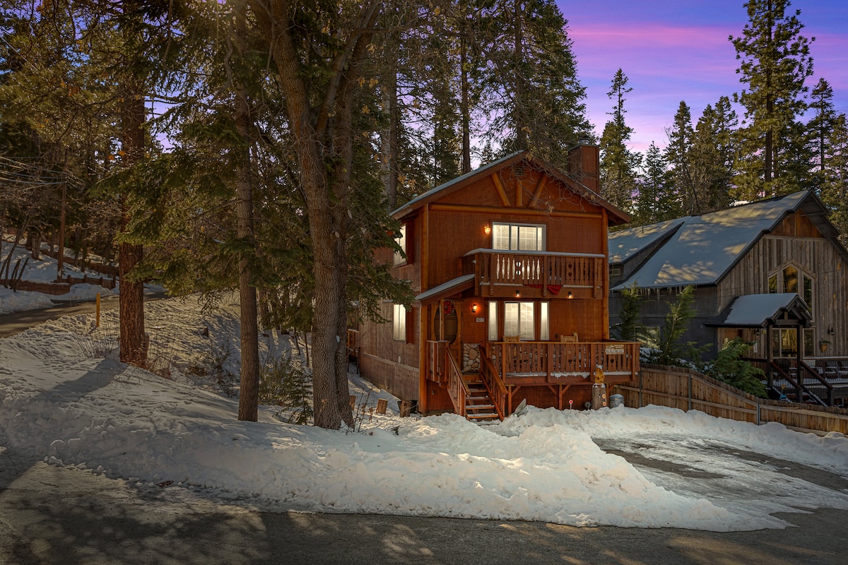 The mid-century modern cabin is shown amidst a snowy landscape, surrounded by tall trees. Warm lighting emanates from the windows, creating a welcoming glow. The two-level structure features a deck, with snow lightly covering the ground and the road visible nearby.