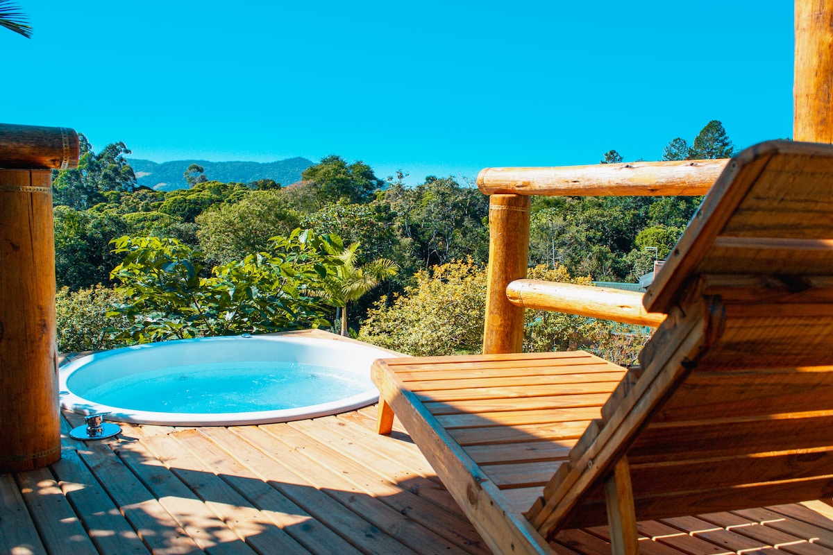 A relaxing outdoor area features a wooden deck with a lounger beside a round hot tub. Lush greenery and trees are visible in the background, complementing the serene environment.