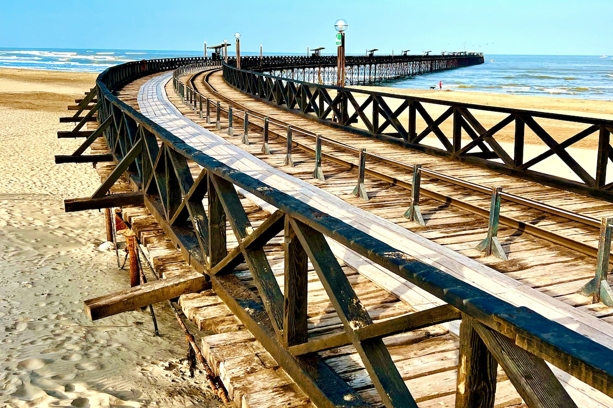 A wooden boardwalk extends along the sandy beach, curving towards a distant pier. The sunlit path is framed by wooden railings, with the ocean visibly stretching beyond. Soft waves gently lap against the shore, creating a serene coastal scene.