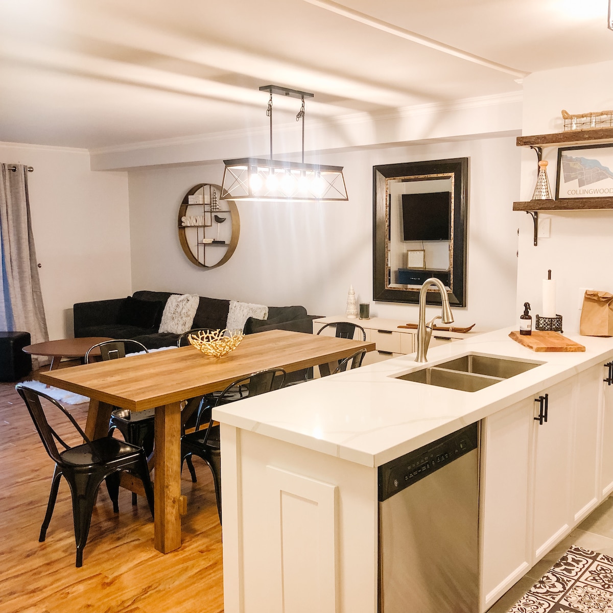The open-concept living area features a spacious dining table with black chairs and a cozy seating arrangement. A modern chandelier hangs above the table, while a large mirror reflects the natural light. A stylish kitchen, with a double sink and sleek countertops, complements the design.
