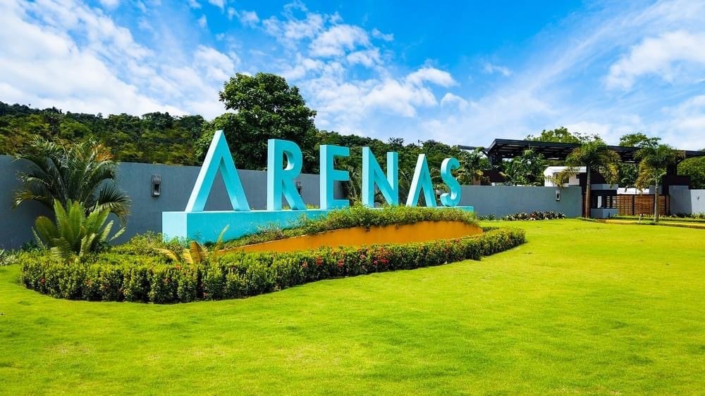 The welcoming entrance to Condominio Arenas features a large, prominently displayed sign against a backdrop of green landscaping. Lush grass and palm plants frame the entrance, while the clear blue sky enhances the inviting atmosphere of the community.