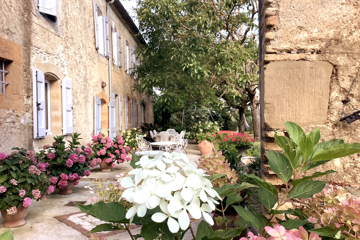 A charming outdoor area is surrounded by lush greenery and flowering plants, with white and pink hydrangeas in prominent display. A patio space is furnished with a white table and chairs, framed by stone walls and a tree, creating a serene atmosphere.