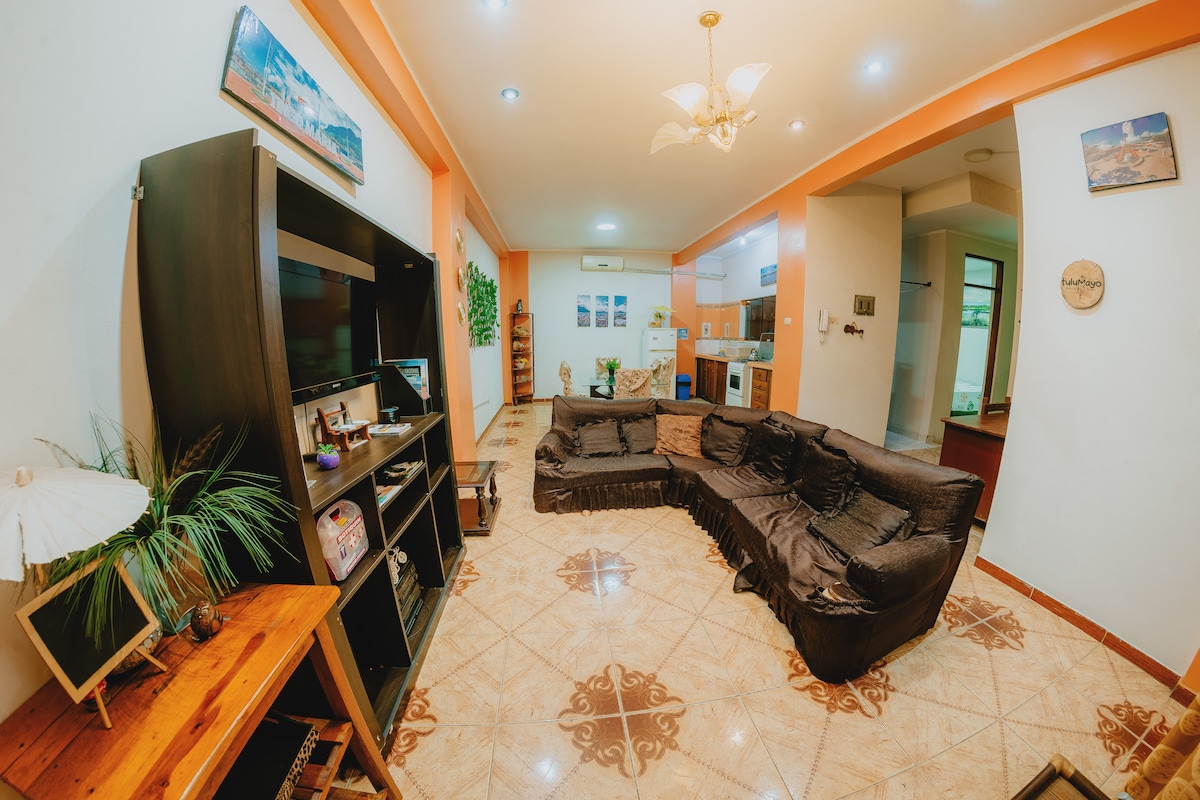 A spacious common area features a large black sectional sofa, providing ample seating. A wooden shelf is seen with decorative items, and plants add a touch of greenery. The room is well-lit, with an open layout leading toward the kitchen area in the background.