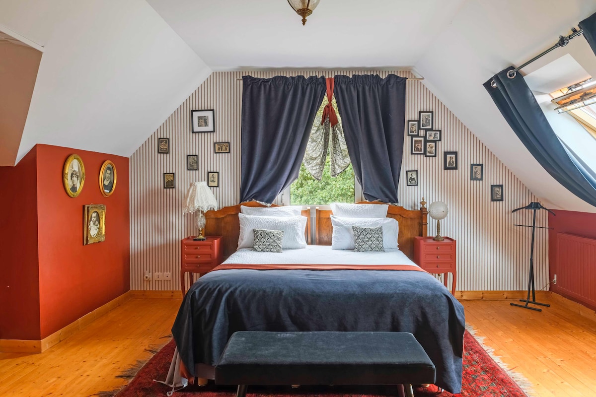 A spacious bedroom features a queen-size bed with soft bedding, complemented by decorative pillows. Vintage black-and-white photographs adorn the walls, while rich red and striped wallpaper adds character. Natural light filters through slanted windows, accented by dark drapes. A small bench sits at the foot of the bed.