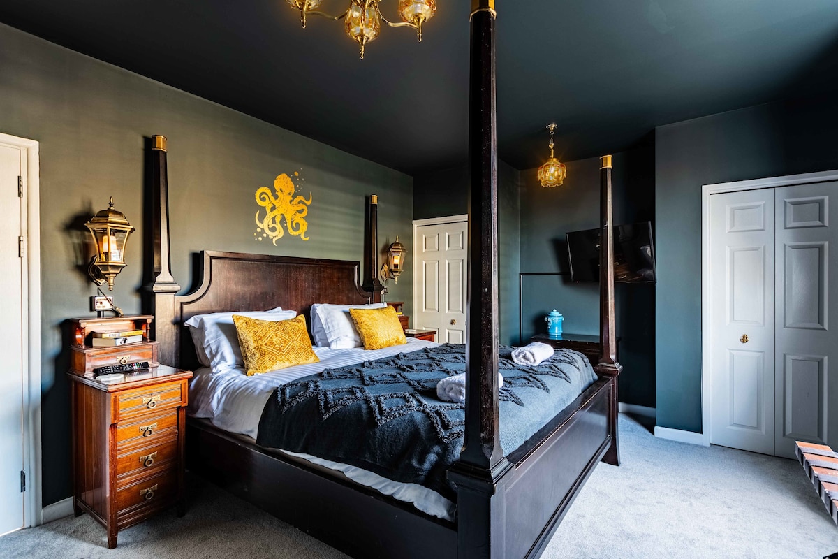 A spacious bedroom features a super king-size four-poster bed adorned with decorative pillows. Wall sconces and a golden octopus motif creatively accent the dark walls. A flat-screen TV is mounted opposite the bed, and vintage furniture adds character to the cozy setting.