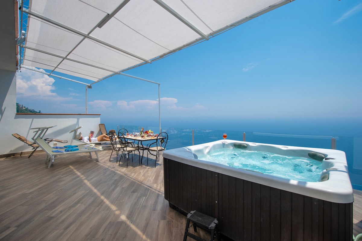 A spacious terrace boasts a hot tub with clear views of the ocean. An outdoor dining set is arranged for six, accompanied by sun loungers. A large awning provides shade, while the blue sky and distant coastline create a serene backdrop for relaxation.