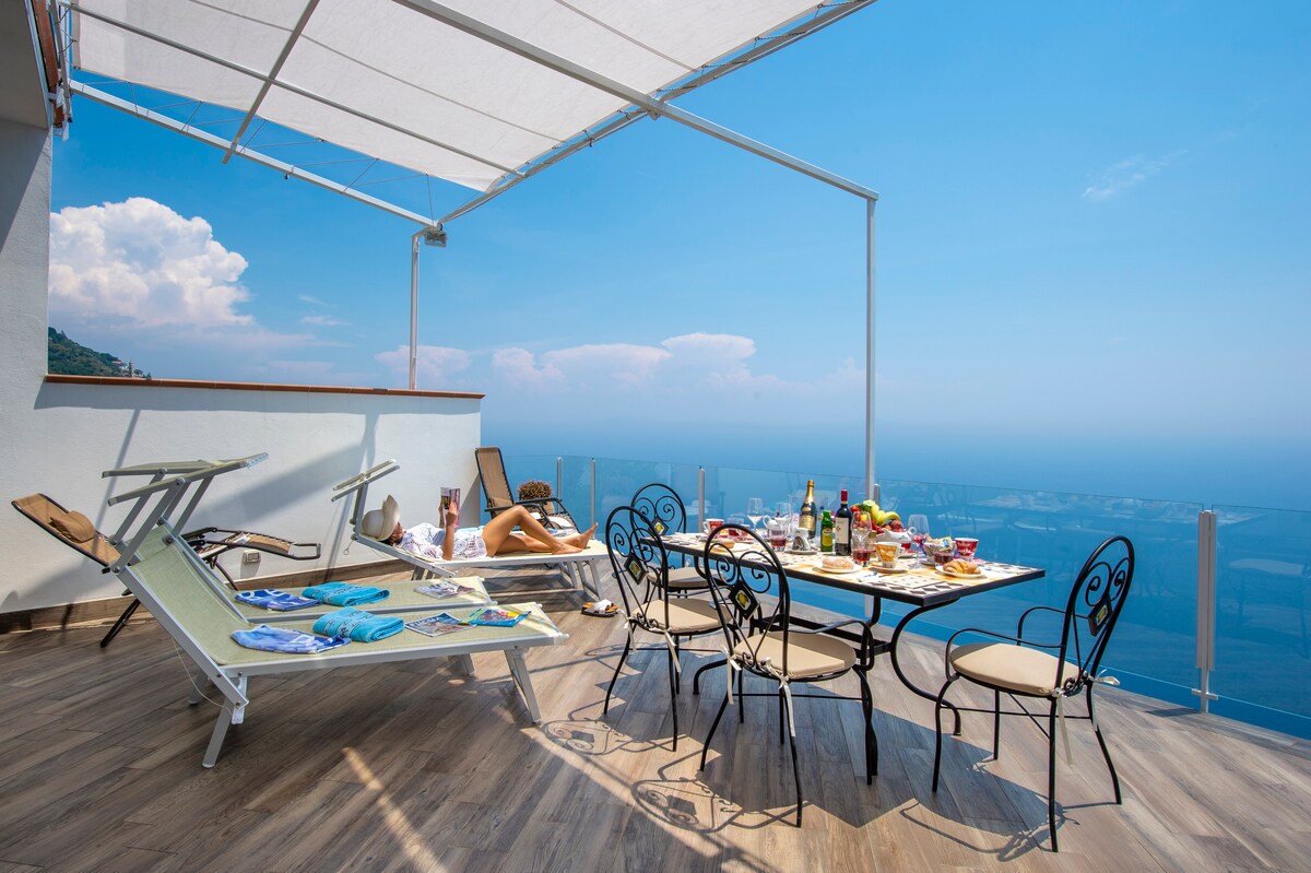 A spacious terrace is furnished with a dining table surrounded by six chairs. Sun loungers are positioned nearby, and a scenic panoramic view can be seen in the background. The table is set with various drinks and snacks, creating an inviting setting for relaxation.