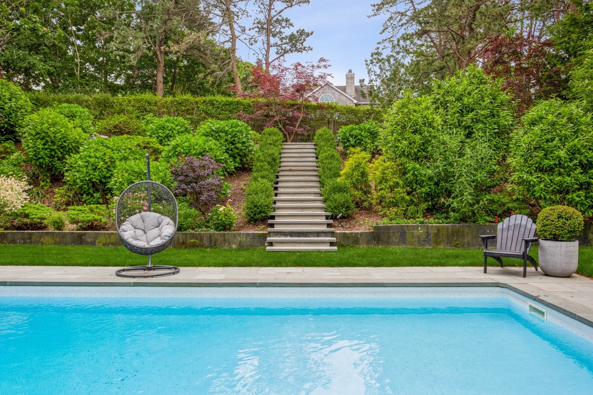 A tranquil pool area features a sparkling blue pool framed by a meticulously landscaped garden. Steps lead up to lush greenery and vibrant flowering plants, while a hanging chair adds a cozy seating option next to the water.