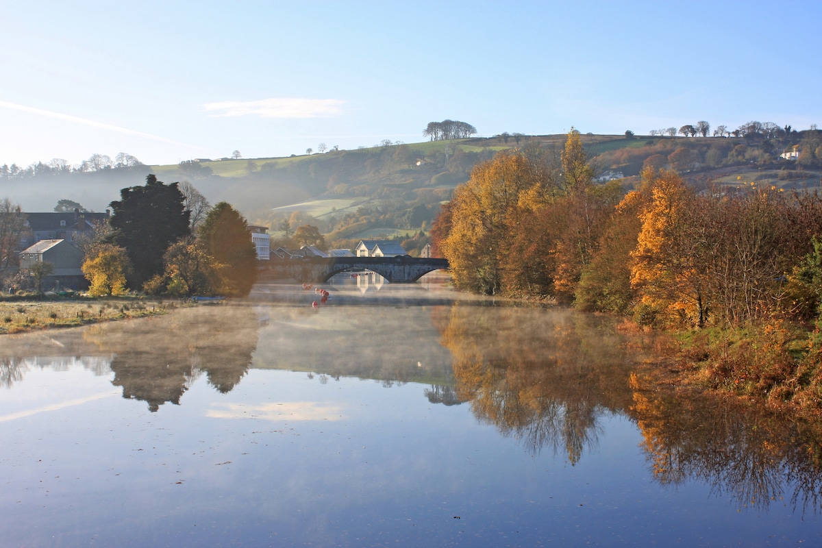 A serene river scene is depicted, featuring calm waters reflecting vibrant autumn trees and a distant bridge. Low morning mist lightly blankets the surface, while the surrounding hills showcase a palette of rich greens and yellows. Clear blue skies provide a tranquil backdrop.