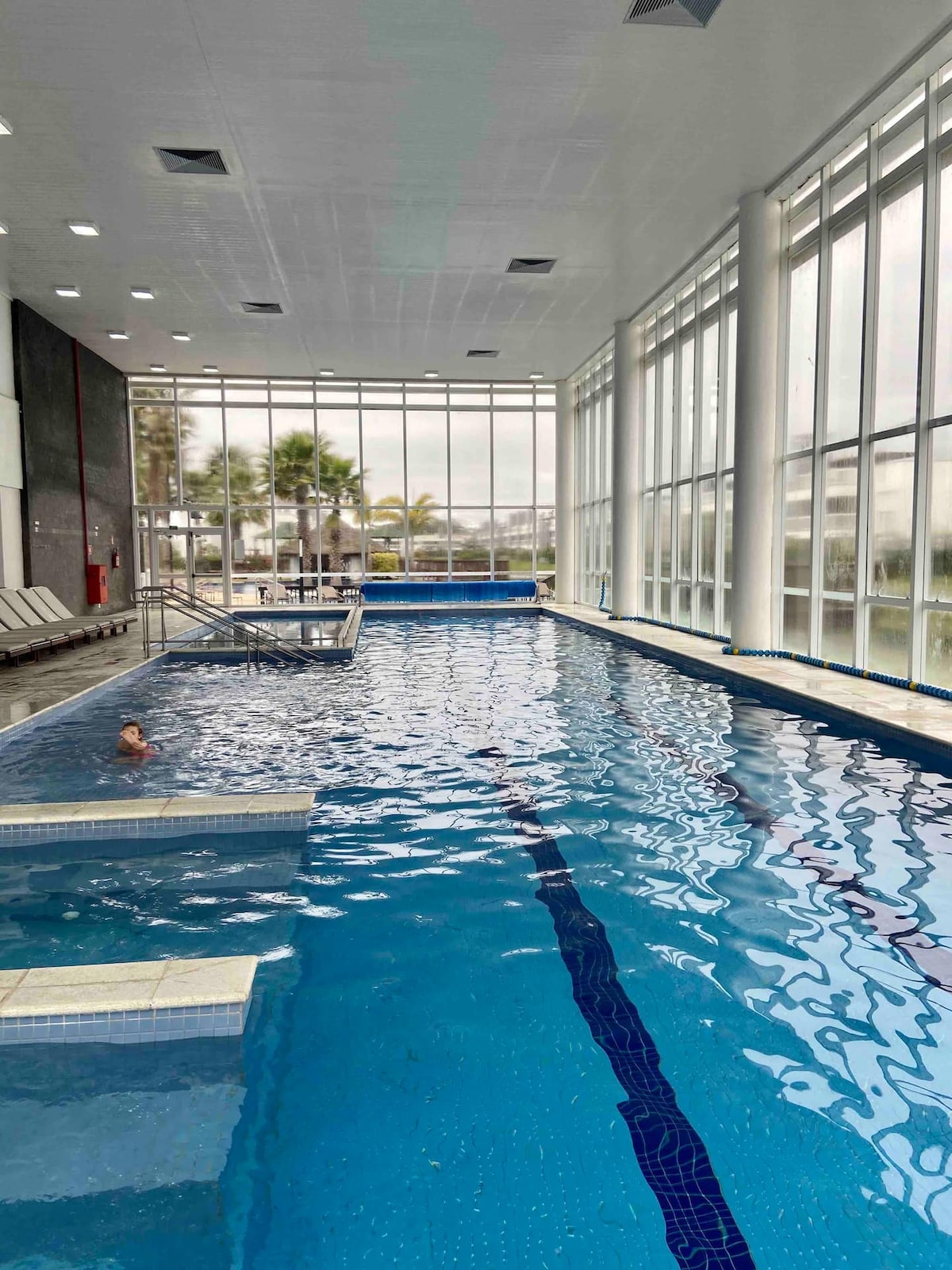 An indoor swimming pool is filled with clear blue water, illuminated by natural light streaming through large windows. Comfortable lounge chairs line one side, while a diving board is positioned near the pool's edge. The atmosphere is spacious and tranquil.