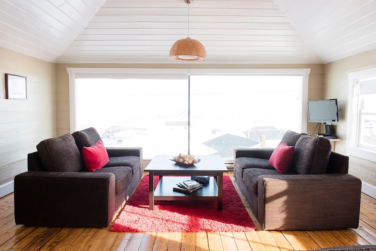 A cozy living area features two dark sofas with red accent cushions facing a central wooden coffee table. A vibrant red rug anchors the space, while large windows provide a view of the surroundings, allowing for ample natural light.