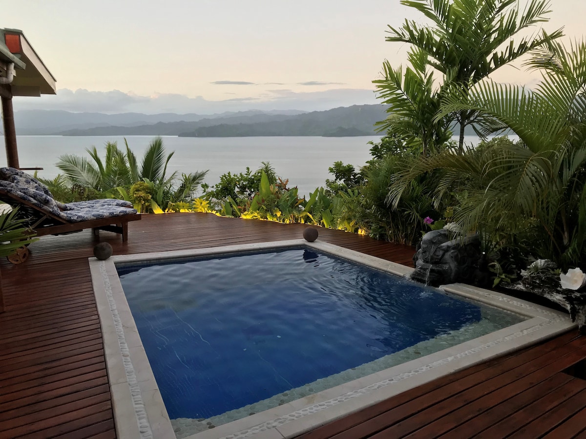 A private hot tub is positioned on a wooden deck, surrounded by lush greenery. Scenic views of the water and distant mountains are visible in the background. Soft lighting enhances the tranquil ambiance as the late afternoon sun casts a warm glow over the area.