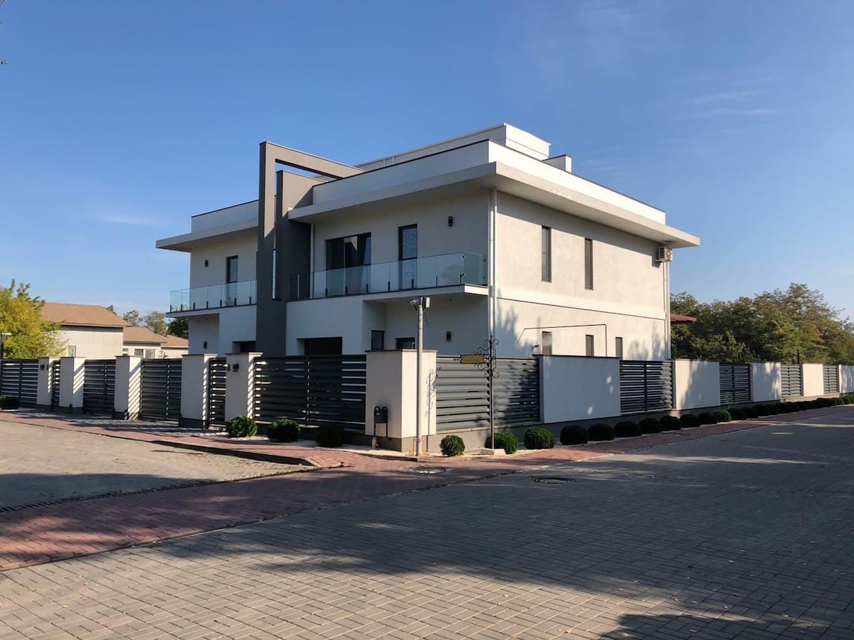 A modern three-level house is displayed, featuring clean lines and a light-colored façade. The property is surrounded by a sleek fence with manicured landscaping and a paved driveway. Balconies are visible on the upper levels, offering outdoor space.