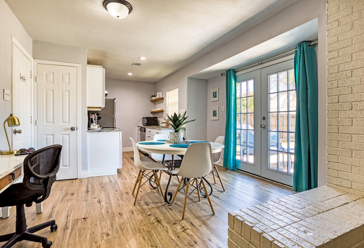 A modern dining area features a circular table surrounded by white chairs, complemented by a large window with turquoise curtains. The space connects to a well-equipped kitchen with light cabinetry, and a door leads to an outdoor area, enhancing the open atmosphere.