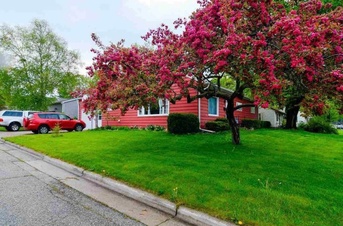 A vintage 1940’s home is set on a landscaped lawn, framed by flowering pink trees. A spacious driveway accommodates multiple vehicles, with a garage visible to the left. The exterior showcases a charming red facade and large front windows, inviting light into the interior.
