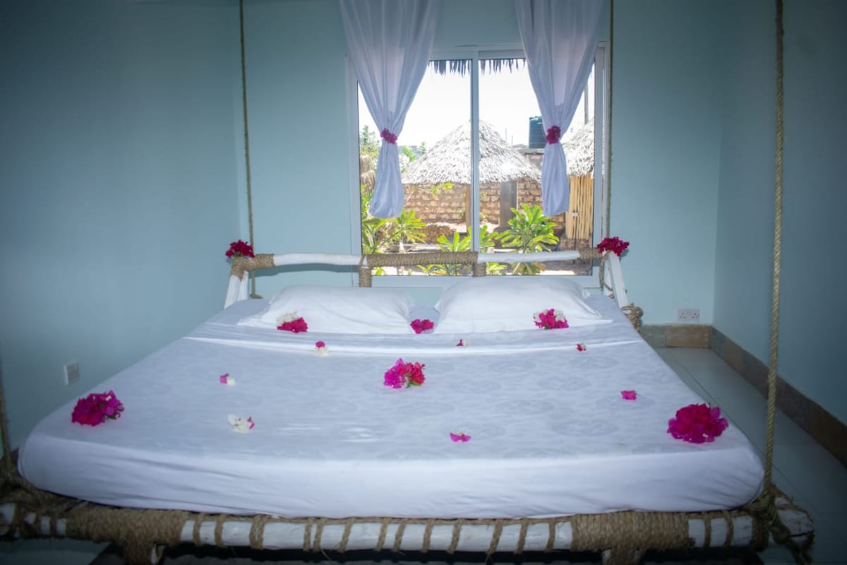 A spacious bed adorned with delicate flower petals is positioned in a bright room. Light enters through large windows, revealing lush greenery outside. Sheer white curtains frame the window, adding a soft touch to the tranquil space.