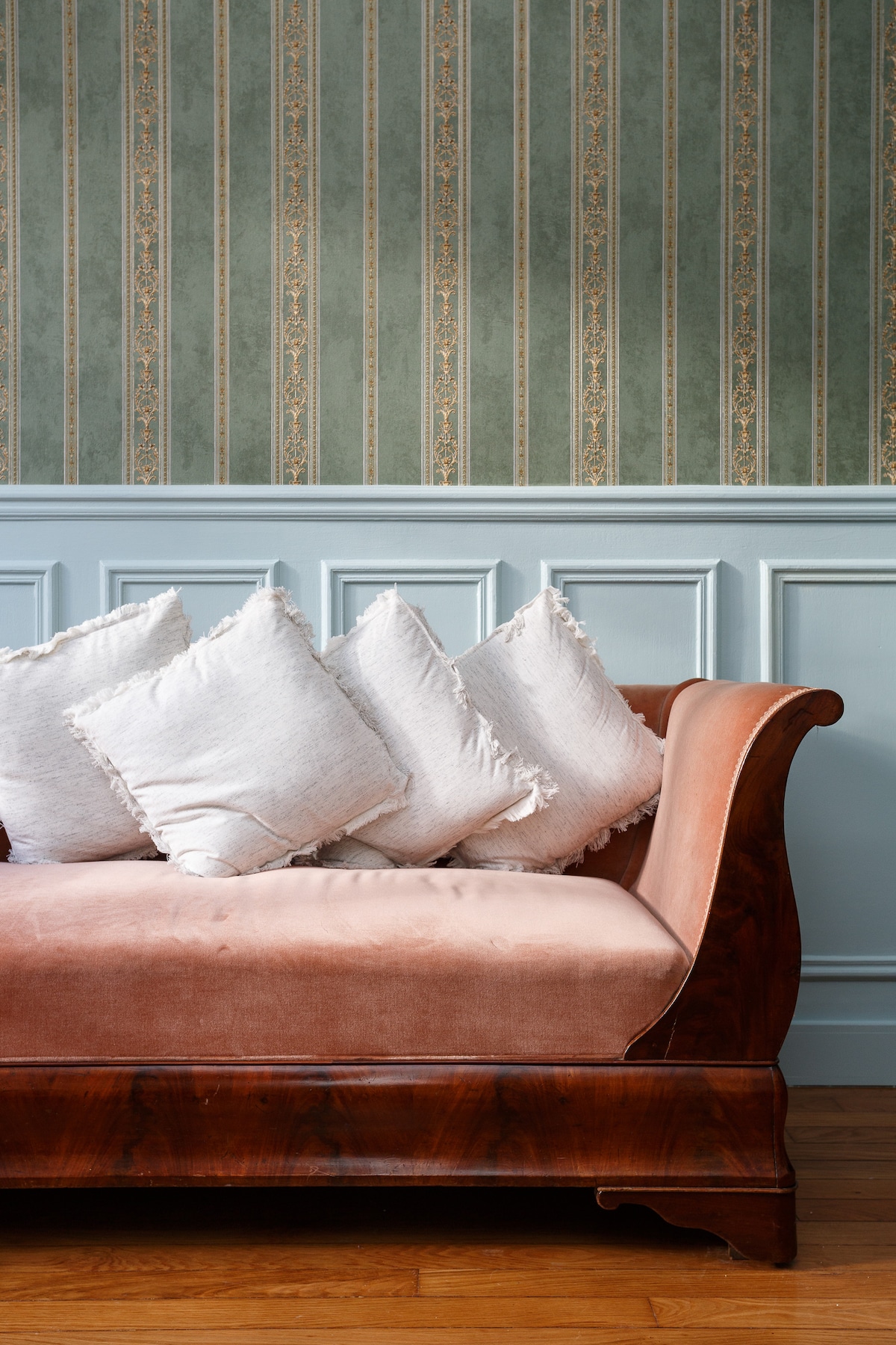A vintage-style sofa with a polished wooden frame is showcased. Soft, frayed cushions are arranged on the seat, complementing the elegant wallpaper with subtle patterns in green and gold. The wainscoting beneath adds a classic touch to the overall ambiance.